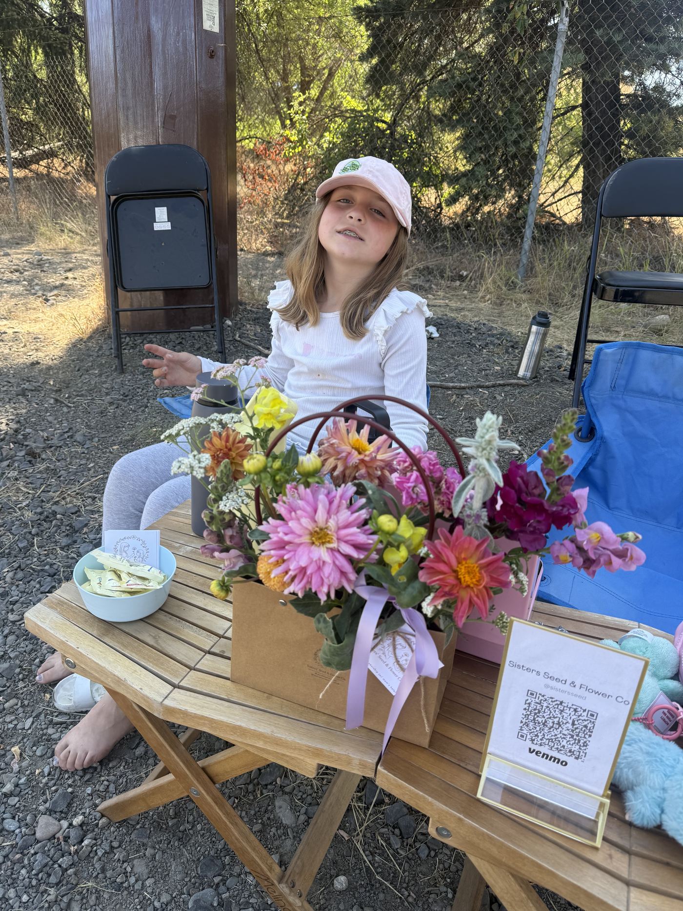 A young girl in a pink cap sitting behind a wooden table covered in a basket of colorful dahlias, snapdragons, and yarrow, with a Sisters Seed & Flower Co. sign and Venmo QR code.