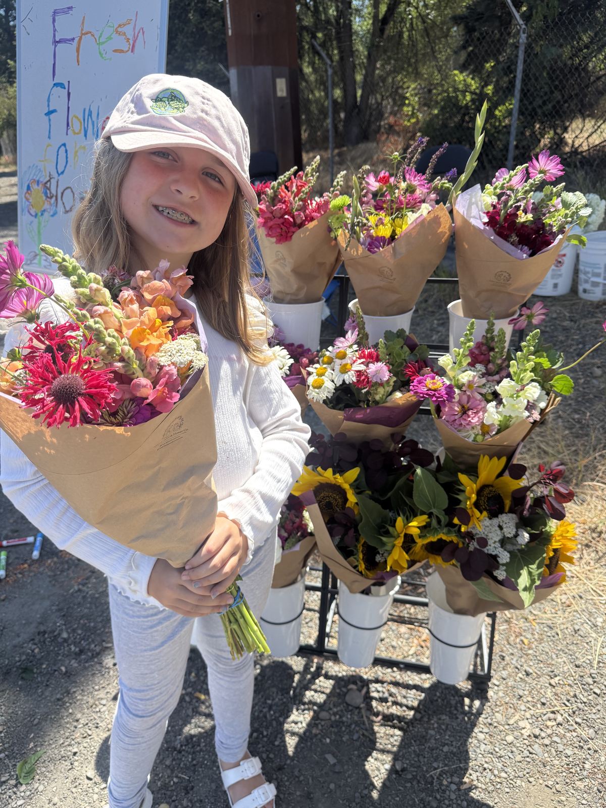 A girl in a pink cap smiling and holding a fresh-cut bouquet of snapdragons, cosmos, and coneflowers in front of the flower stand.