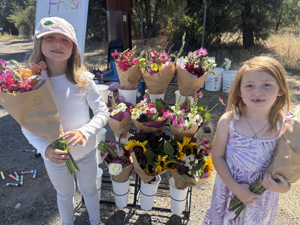 Two sisters standing together at the roadside flower stand, each holding freshly cut bouquets, with the Fresh Flowers for Sale sign behind them.